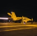 A U.S. Navy carrier-based jet aircraft, numbered 504, is preparing for a night launch on an aircraft carrier flight deck. The aircraft's afterburner is visibly ignited, producing a bright exhaust plume to the left. Multiple flight deck crew members in full gear are positioned around the aircraft observing or managing the launch sequence.