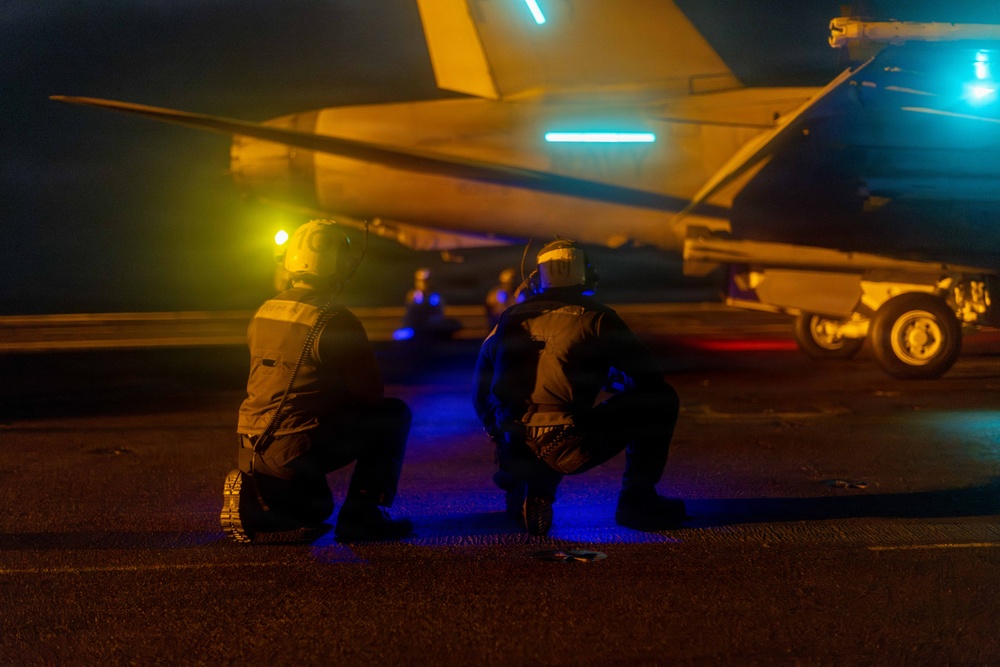Two military deck crew personnel are kneeling on what appears to be an aircraft carrier flight deck at night, positioned near a military jet aircraft. The scene is illuminated by colored lights including yellow, blue, and cyan, which are characteristic of carrier flight deck operations. The personnel are wearing flight deck gear including helmets with integrated lighting.