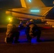 Two military deck crew personnel are kneeling on what appears to be an aircraft carrier flight deck at night, positioned near a military jet aircraft. The scene is illuminated by colored lights including yellow, blue, and cyan, which are characteristic of carrier flight deck operations. The personnel are wearing flight deck gear including helmets with integrated lighting.