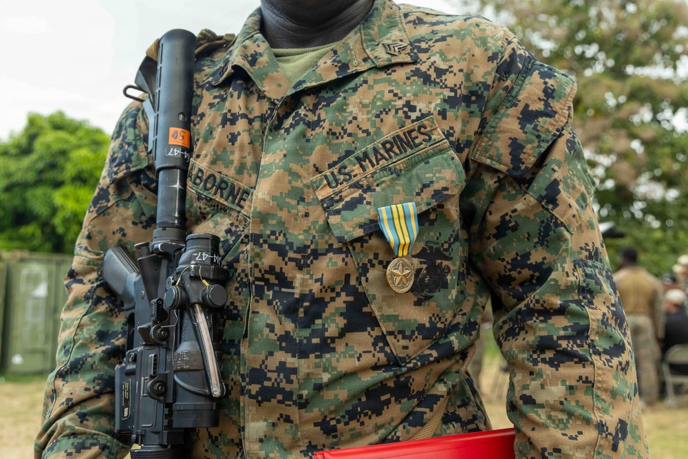 A U.S. Marine is shown from the neck down wearing Marine Pattern (MARPAT) woodland digital camouflage uniform. The Marine is carrying a rifle with a scope and has a service medal pinned to their chest. The name tape reads 'ABORNE' and the branch tape reads 'U.S. MARINES'.