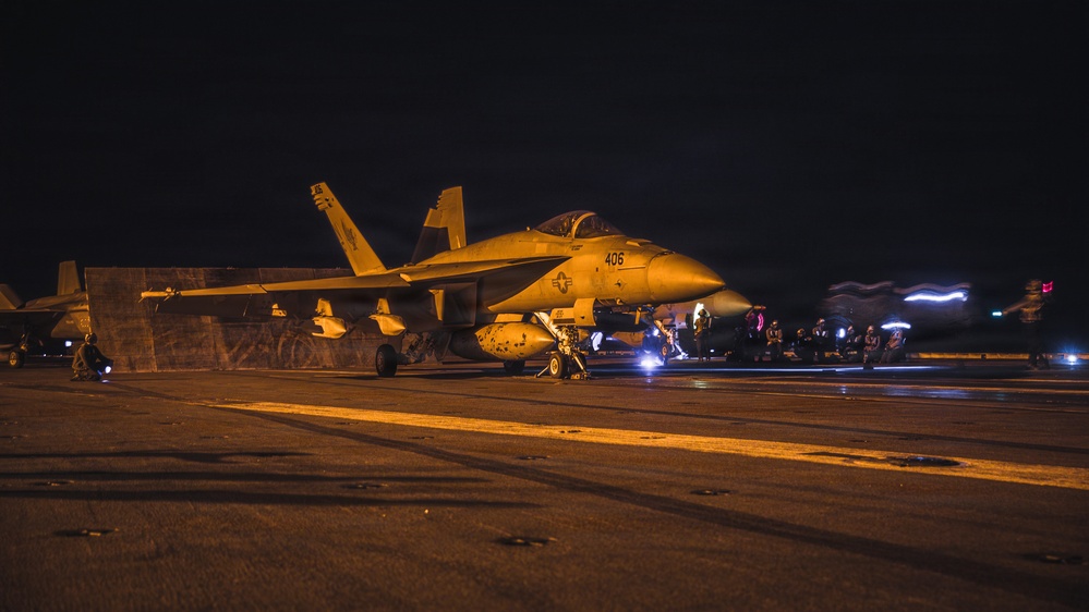 A U.S. Navy carrier-based jet aircraft, marked '406', is positioned on what appears to be an aircraft carrier flight deck during nighttime operations. Ground crew personnel are visible around the aircraft conducting pre-flight or post-flight procedures. The scene is illuminated by warm artificial lighting, creating an orange-tinted atmosphere against a dark sky.
