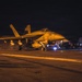 A U.S. Navy carrier-based jet aircraft, marked '406', is positioned on what appears to be an aircraft carrier flight deck during nighttime operations. Ground crew personnel are visible around the aircraft conducting pre-flight or post-flight procedures. The scene is illuminated by warm artificial lighting, creating an orange-tinted atmosphere against a dark sky.