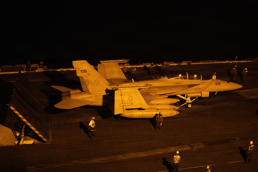 A U.S. Navy F/A-18 Hornet or Super Hornet (bureau number 106) is positioned on an aircraft carrier flight deck at night, illuminated by warm orange artificial lighting. Multiple flight deck personnel in various colored jerseys are visible around the aircraft, suggesting pre-launch or post-recovery operations. The scene depicts a typical nighttime carrier flight deck environment with characteristic low-light conditions.