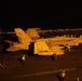 A U.S. Navy F/A-18 Hornet or Super Hornet (bureau number 106) is positioned on an aircraft carrier flight deck at night, illuminated by warm orange artificial lighting. Multiple flight deck personnel in various colored jerseys are visible around the aircraft, suggesting pre-launch or post-recovery operations. The scene depicts a typical nighttime carrier flight deck environment with characteristic low-light conditions.