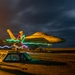 A long-exposure night photograph of a military jet fighter, likely an F/A-18 Hornet or Super Hornet, parked on what appears to be an aircraft carrier flight deck. Colorful light trails in blue, green, and red are visible around the aircraft, created by the movement of crew members carrying light wands or flashlights during the long exposure. The dramatic dark sky with clouds and warm deck lighting create a striking visual composition.