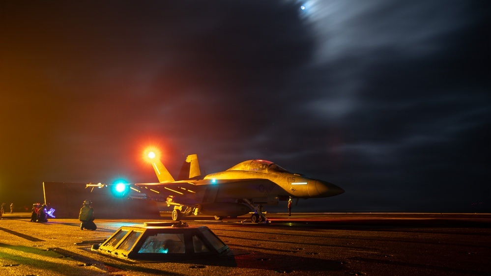 A military jet aircraft, consistent with an F/A-18 Hornet or Super Hornet, is positioned on what appears to be an aircraft carrier flight deck at night. The scene is illuminated by colored signal lights (red and green/teal) and ambient orange lighting, with deck crew personnel visible around the aircraft. The dramatic night sky with cloud formations and a light streak suggests active flight operations.