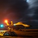 A military jet aircraft, consistent with an F/A-18 Hornet or Super Hornet, is positioned on what appears to be an aircraft carrier flight deck at night. The scene is illuminated by colored signal lights (red and green/teal) and ambient orange lighting, with deck crew personnel visible around the aircraft. The dramatic night sky with cloud formations and a light streak suggests active flight operations.