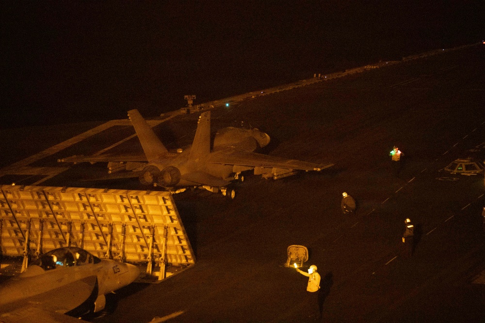 A nighttime scene on what appears to be a naval aircraft carrier flight deck, showing a military jet aircraft (likely an F/A-18 Hornet or Super Hornet) positioned near the deck edge elevator. Flight deck crew members are visible using illuminated wands to direct aircraft operations. The scene is lit primarily by artificial lighting, creating strong contrast against the dark surroundings.
