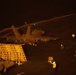 A nighttime scene on what appears to be a naval aircraft carrier flight deck, showing a military jet aircraft (likely an F/A-18 Hornet or Super Hornet) positioned near the deck edge elevator. Flight deck crew members are visible using illuminated wands to direct aircraft operations. The scene is lit primarily by artificial lighting, creating strong contrast against the dark surroundings.
