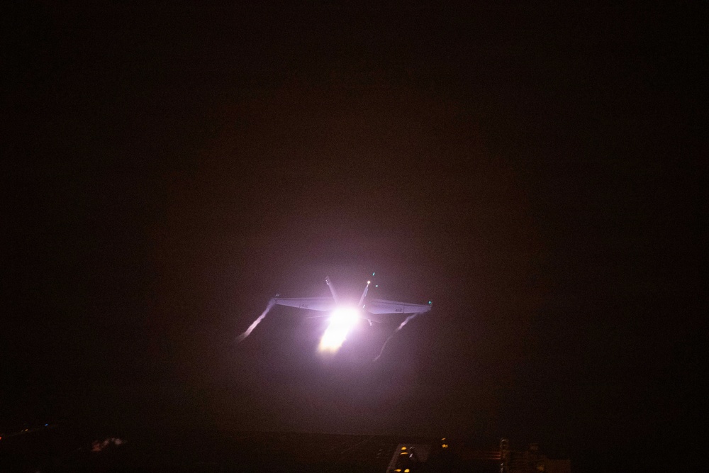 A nighttime photograph showing an aircraft or aerial object illuminated by a bright light source, possibly afterburners or landing/navigation lights. The image is taken in very low-light conditions, making fine details difficult to discern. Some figures or structures are faintly visible at the bottom edge of the frame.