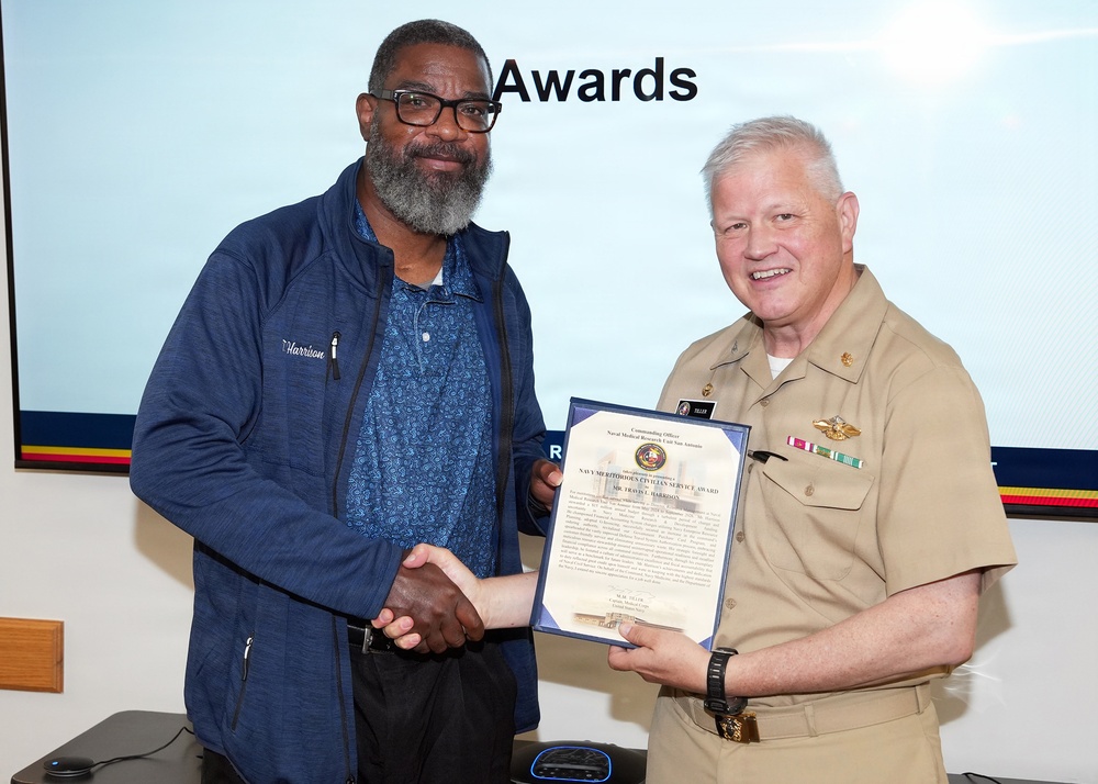 A U.S. Navy officer in tan service uniform is presenting a Navy Meritorious Civilian Service Award certificate to a civilian man named Harrison during a formal awards ceremony. The two individuals are shaking hands while jointly holding the framed certificate. The event appears to be taking place in a conference or briefing room with a presentation screen displaying the word 'Awards' in the background.
