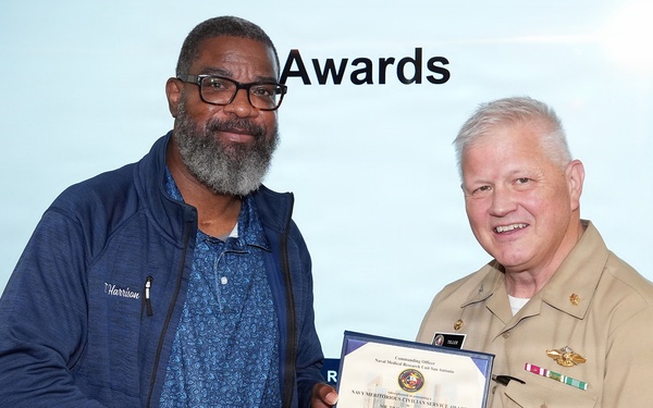 A U.S. Navy officer in tan service uniform is presenting a Navy Meritorious Civilian Service Award certificate to a civilian man named Harrison during a formal awards ceremony. The two individuals are shaking hands while jointly holding the framed certificate. The event appears to be taking place in a conference or briefing room with a presentation screen displaying the word 'Awards' in the background.