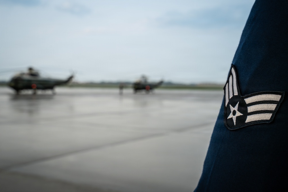 A close-up photograph focusing on the sleeve insignia of a United States Air Force Airman First Class uniform in the foreground, with two military helicopters visible on a wet tarmac in the blurred background. The rank patch shows the distinctive chevron and star design of an Airman First Class (E-3). The setting appears to be a military airfield under overcast conditions.