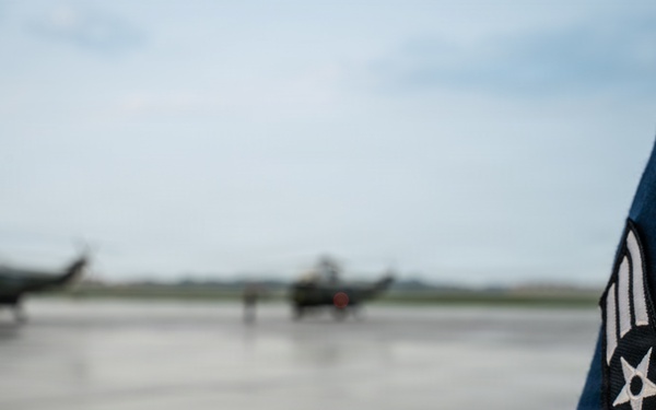 A close-up photograph focusing on the sleeve insignia of a United States Air Force Airman First Class uniform in the foreground, with two military helicopters visible on a wet tarmac in the blurred background. The rank patch shows the distinctive chevron and star design of an Airman First Class (E-3). The setting appears to be a military airfield under overcast conditions.