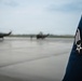 A close-up photograph focusing on the sleeve insignia of a United States Air Force Airman First Class uniform in the foreground, with two military helicopters visible on a wet tarmac in the blurred background. The rank patch shows the distinctive chevron and star design of an Airman First Class (E-3). The setting appears to be a military airfield under overcast conditions.
