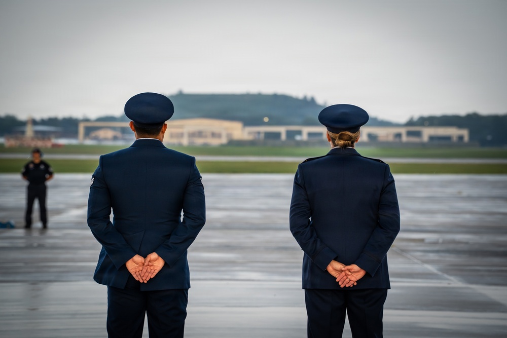 Two military personnel in dress blue uniforms stand at parade rest on a wet airfield tarmac, facing away from the camera toward the runway. A third individual in dark clothing is visible in the background at a distance. The setting appears to be a formal military ceremony or dignitary arrival at an air base.