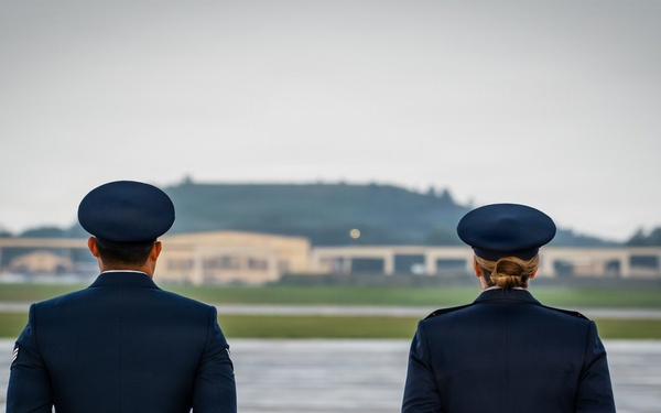 Two military personnel in dress blue uniforms stand at parade rest on a wet airfield tarmac, facing away from the camera toward the runway. A third individual in dark clothing is visible in the background at a distance. The setting appears to be a formal military ceremony or dignitary arrival at an air base.