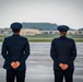 Two military personnel in dress blue uniforms stand at parade rest on a wet airfield tarmac, facing away from the camera toward the runway. A third individual in dark clothing is visible in the background at a distance. The setting appears to be a formal military ceremony or dignitary arrival at an air base.