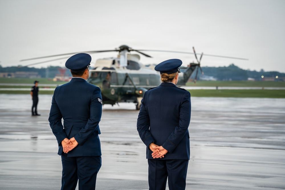 Two uniformed military personnel, standing at parade rest with hands clasped behind their backs, face a large military helicopter on a wet tarmac. The scene appears to be a formal arrival or departure ceremony at a military airfield. Overcast skies and a reflective wet runway surface suggest recent rainfall.