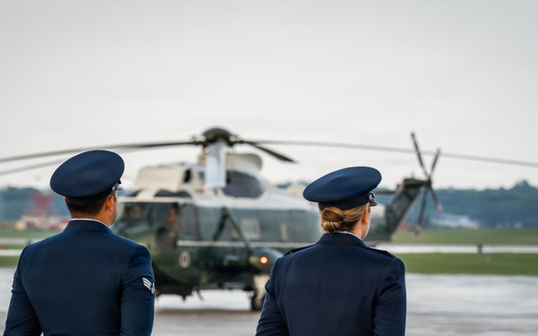 Two uniformed military personnel, standing at parade rest with hands clasped behind their backs, face a large military helicopter on a wet tarmac. The scene appears to be a formal arrival or departure ceremony at a military airfield. Overcast skies and a reflective wet runway surface suggest recent rainfall.