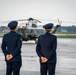 Two uniformed military personnel, standing at parade rest with hands clasped behind their backs, face a large military helicopter on a wet tarmac. The scene appears to be a formal arrival or departure ceremony at a military airfield. Overcast skies and a reflective wet runway surface suggest recent rainfall.
