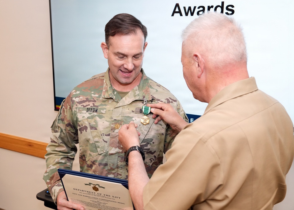 A military awards ceremony is taking place where a person in a tan/khaki uniform is pinning a medal onto a soldier in Army OCP (Operational Camouflage Pattern) uniform with the name tape 'DIXON'. The soldier is holding a Department of the Navy certificate identifying the award as the Navy and Marine Corps Commendation Medal. The word 'Awards' is visible on a presentation screen in the background.