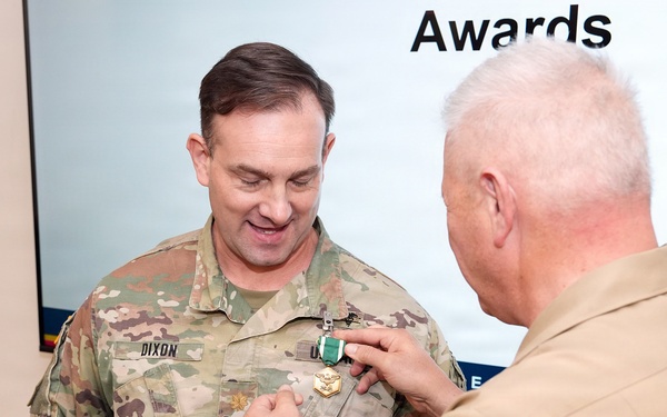 A military awards ceremony is taking place where a person in a tan/khaki uniform is pinning a medal onto a soldier in Army OCP (Operational Camouflage Pattern) uniform with the name tape 'DIXON'. The soldier is holding a Department of the Navy certificate identifying the award as the Navy and Marine Corps Commendation Medal. The word 'Awards' is visible on a presentation screen in the background.