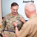 A military awards ceremony is taking place where a person in a tan/khaki uniform is pinning a medal onto a soldier in Army OCP (Operational Camouflage Pattern) uniform with the name tape 'DIXON'. The soldier is holding a Department of the Navy certificate identifying the award as the Navy and Marine Corps Commendation Medal. The word 'Awards' is visible on a presentation screen in the background.