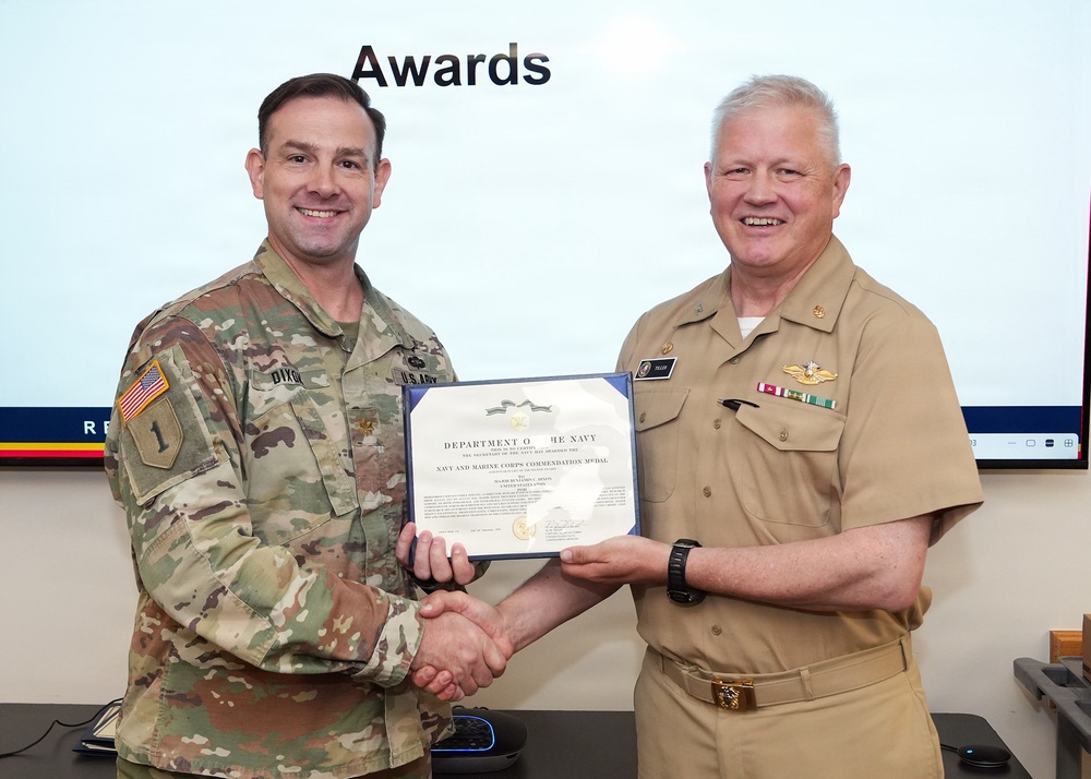 Two military officers are shaking hands while jointly holding an award certificate during a formal awards ceremony. The certificate is from the Department of the Navy and appears to be a Navy and Marine Corps Commendation Medal. One officer is in U.S. Army combat uniform and the other is in U.S. Navy service khaki uniform.