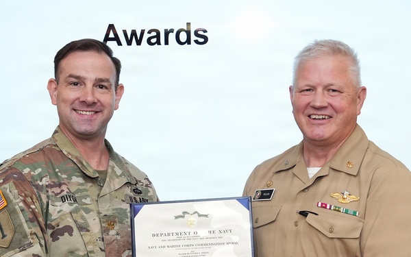Two military officers are shaking hands while jointly holding an award certificate during a formal awards ceremony. The certificate is from the Department of the Navy and appears to be a Navy and Marine Corps Commendation Medal. One officer is in U.S. Army combat uniform and the other is in U.S. Navy service khaki uniform.