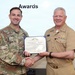 Two military officers are shaking hands while jointly holding an award certificate during a formal awards ceremony. The certificate is from the Department of the Navy and appears to be a Navy and Marine Corps Commendation Medal. One officer is in U.S. Army combat uniform and the other is in U.S. Navy service khaki uniform.