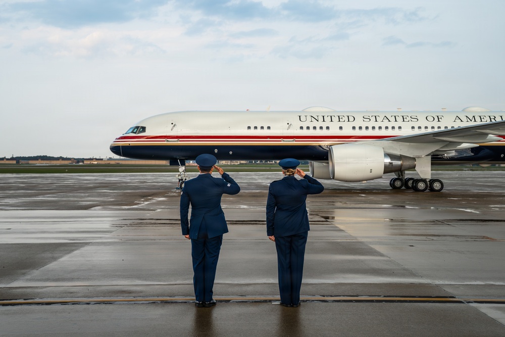 Two U.S. Air Force personnel in dress blue uniforms are standing on a wet tarmac, rendering a salute toward a U.S. government aircraft bearing the inscription 'United States of America.' The aircraft features a distinctive livery with a white fuselage, gold lettering, and red and dark blue accent stripes, consistent with U.S. Air Force executive transport aircraft. The scene appears to be a formal arrival or departure ceremony at a military airfield.