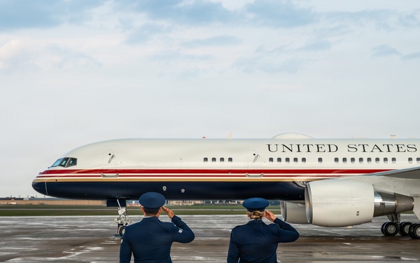Two U.S. Air Force personnel in dress blue uniforms are standing on a wet tarmac, rendering a salute toward a U.S. government aircraft bearing the inscription 'United States of America.' The aircraft features a distinctive livery with a white fuselage, gold lettering, and red and dark blue accent stripes, consistent with U.S. Air Force executive transport aircraft. The scene appears to be a formal arrival or departure ceremony at a military airfield.