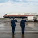 Two U.S. Air Force personnel in dress blue uniforms are standing on a wet tarmac, rendering a salute toward a U.S. government aircraft bearing the inscription 'United States of America.' The aircraft features a distinctive livery with a white fuselage, gold lettering, and red and dark blue accent stripes, consistent with U.S. Air Force executive transport aircraft. The scene appears to be a formal arrival or departure ceremony at a military airfield.