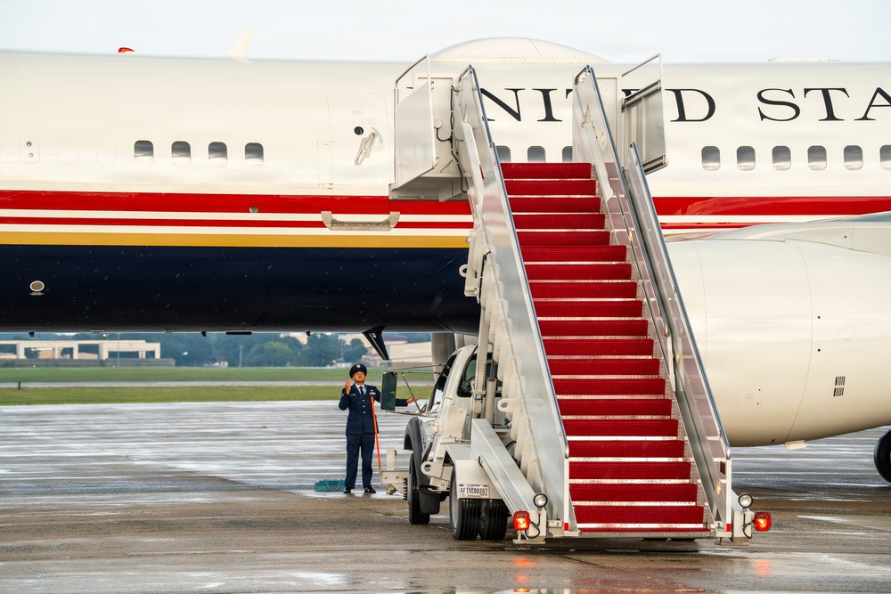 A large United States government aircraft is parked on a wet tarmac with a red-carpeted mobile staircase positioned at the door. A uniformed military personnel member stands at attention near the base of the stairs. The aircraft displays distinctive red, white, and blue livery with 'UNITED STATES' lettering visible on the fuselage.
