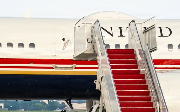 A large United States government aircraft is parked on a wet tarmac with a red-carpeted mobile staircase positioned at the door. A uniformed military personnel member stands at attention near the base of the stairs. The aircraft displays distinctive red, white, and blue livery with 'UNITED STATES' lettering visible on the fuselage.