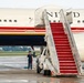A large United States government aircraft is parked on a wet tarmac with a red-carpeted mobile staircase positioned at the door. A uniformed military personnel member stands at attention near the base of the stairs. The aircraft displays distinctive red, white, and blue livery with 'UNITED STATES' lettering visible on the fuselage.