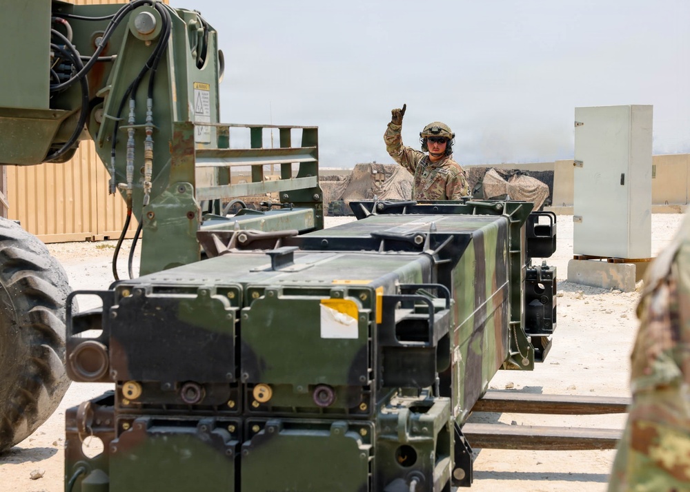 A military service member in combat uniform and helmet is giving a hand signal, likely directing a loading or positioning operation. The scene involves military equipment including what appears to be missile launcher pods or ammunition containers being handled by a forklift or loader vehicle. The setting appears to be a forward operating base or military installation in an arid environment.