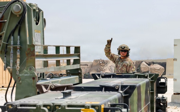 A military service member in combat uniform and helmet is giving a hand signal, likely directing a loading or positioning operation. The scene involves military equipment including what appears to be missile launcher pods or ammunition containers being handled by a forklift or loader vehicle. The setting appears to be a forward operating base or military installation in an arid environment.