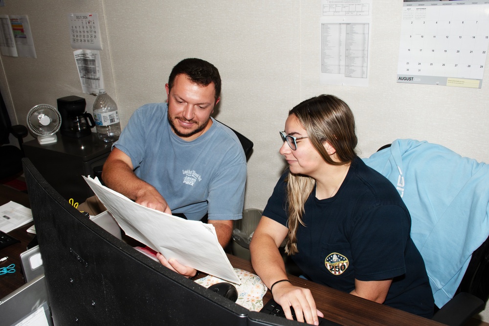 Two individuals are seated at a desk in an office environment, reviewing documents together. A man in a light blue t-shirt and a woman in a dark navy t-shirt with a circular patch logo appear to be collaborating on paperwork. The setting includes office supplies, a small fan, a coffee maker, and wall calendars showing July and August.