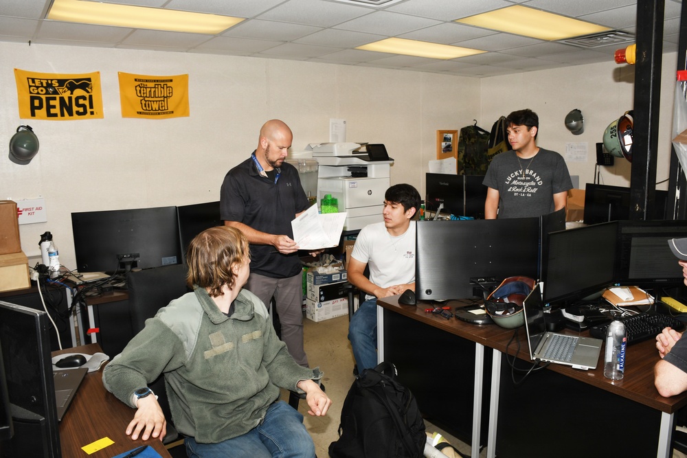 An office or workspace setting with four visible individuals gathered around computer workstations. A bald man in a black polo shirt appears to be reviewing or presenting a document to others. The room contains multiple monitors, laptops, and Pittsburgh Penguins-themed sports banners on the wall.