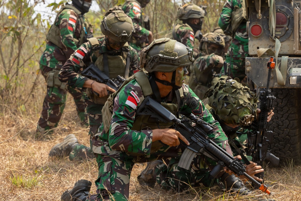 The image shows a group of Indonesian military soldiers conducting what appears to be a tactical field exercise or training operation. Soldiers are positioned in a crouching/low stance near a military vehicle, armed with assault rifles and wearing full combat gear. The setting is a dry, scrubby outdoor terrain consistent with a training environment.