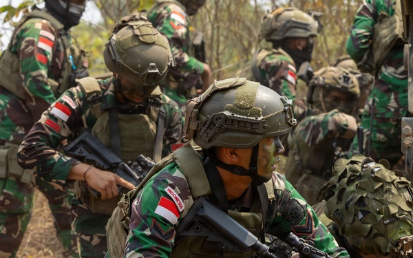 The image shows a group of Indonesian military soldiers conducting what appears to be a tactical field exercise or training operation. Soldiers are positioned in a crouching/low stance near a military vehicle, armed with assault rifles and wearing full combat gear. The setting is a dry, scrubby outdoor terrain consistent with a training environment.
