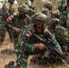 The image shows a group of Indonesian military soldiers conducting what appears to be a tactical field exercise or training operation. Soldiers are positioned in a crouching/low stance near a military vehicle, armed with assault rifles and wearing full combat gear. The setting is a dry, scrubby outdoor terrain consistent with a training environment.