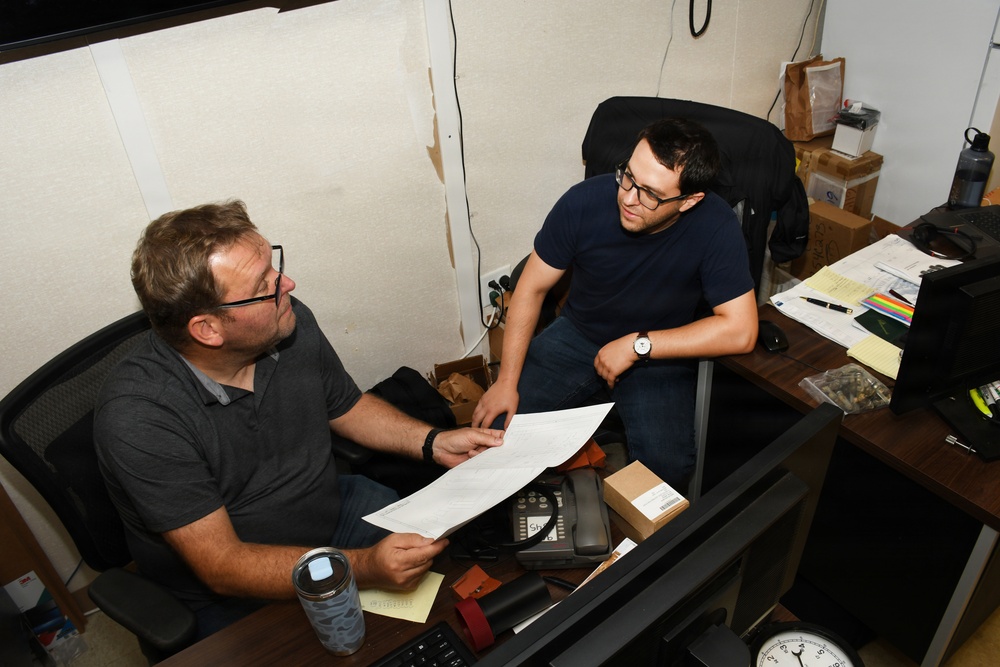 Two men wearing glasses are seated in an office workspace, engaged in a discussion over a document being held by the man on the left. The setting appears to be a cluttered, informal office or workspace with a desk covered in papers, a desk phone, and various items. The interaction suggests a work-related review or consultation.