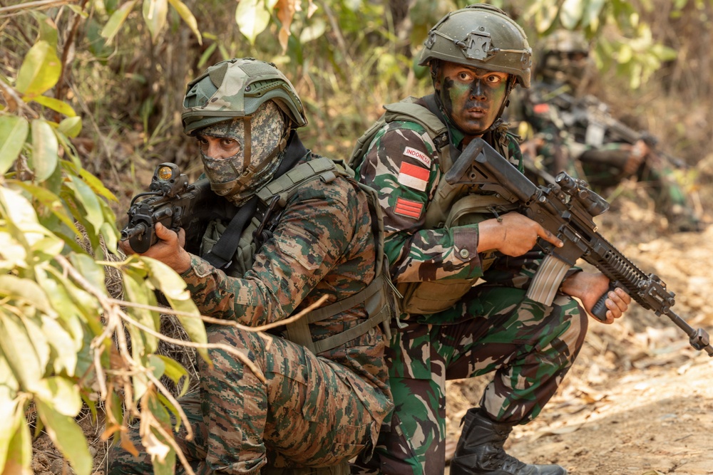 The image shows two armed soldiers in tactical positions in a wooded/brush environment. The soldier on the right wears an 'INDONESIA' patch and the Indonesian flag on their uniform, while the soldier on the left wears a face covering and different camouflage pattern. A third soldier is partially visible in the background, suggesting a small unit tactical exercise or operation.