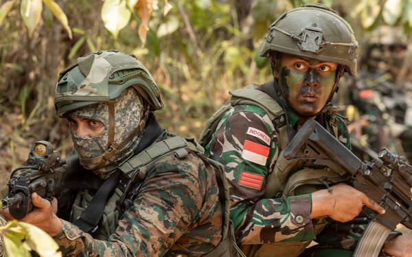 The image shows two armed soldiers in tactical positions in a wooded/brush environment. The soldier on the right wears an 'INDONESIA' patch and the Indonesian flag on their uniform, while the soldier on the left wears a face covering and different camouflage pattern. A third soldier is partially visible in the background, suggesting a small unit tactical exercise or operation.