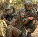 The image shows two armed soldiers in tactical positions in a wooded/brush environment. The soldier on the right wears an 'INDONESIA' patch and the Indonesian flag on their uniform, while the soldier on the left wears a face covering and different camouflage pattern. A third soldier is partially visible in the background, suggesting a small unit tactical exercise or operation.