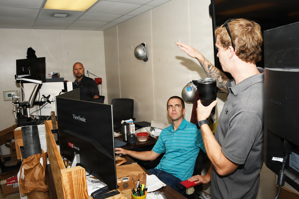 Three men are gathered in a cluttered office workspace. One man sits at a desk using a ViewSonic monitor, while a second man with tattoos and headphones around his neck gestures expressively, appearing to explain something. A third bald man stands in the background observing.
