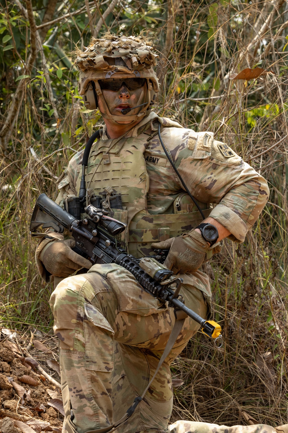 A U.S. Army soldier in full combat gear is positioned in a wooded/brushy outdoor environment. The soldier is wearing MultiCam (OCP) camouflage uniform with a plate carrier vest, tactical helmet with camouflage cover, and face paint. The soldier is holding an M4-style carbine rifle with an optic and appears to be in a field training or operational setting.